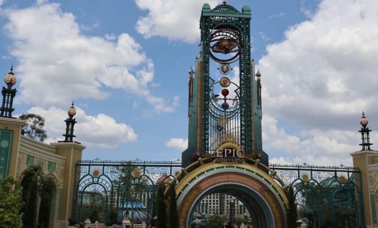 Ornate entrance gate with "Epic Universe" sign, tall decorative tower, and celestial-themed elements under a partly cloudy sky, welcoming guests to the enchanting world of Epic Universe with greenery and people visible beyond as the Universal Epic Universe opening takes place as a new expansion project could be coming.