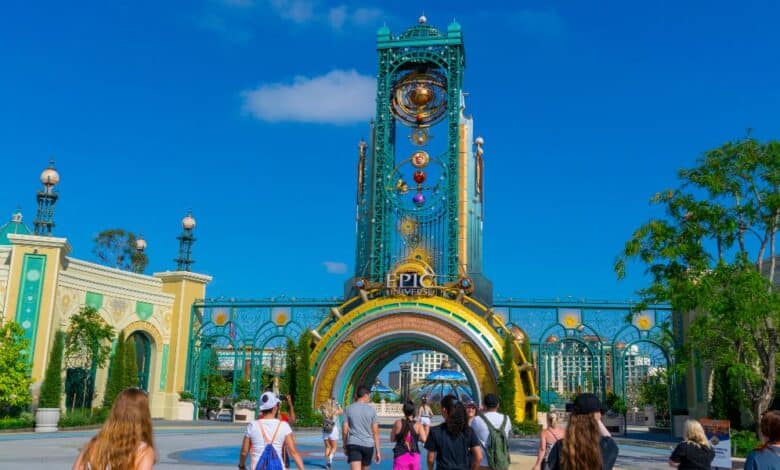 A group of people walk toward the grand, colorful entrance arch and tall clock tower of Universal’s Epic Universe theme park under a bright blue sky. Lush trees and ornate decorations frame the scene with refunds being handed out as Universal Epic Universe preps for its grand-opening.