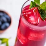 A tall glass of red berry drink with ice cubes, garnished with a sprig of fresh mint, sits on a light surface. A small bowl of blueberries is blurred in the background.