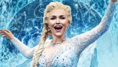 A performer in a sparkling, light blue ice-themed costume with long blond hair styled in a braid smiles and spreads her arms, evoking Frozen the Musical as she stands before an icy, snowflake-patterned backdrop.
