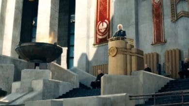 A person stands at a podium on a grand stone platform with burning cauldrons and red banners, addressing seated officials outside a large, imposing building.
