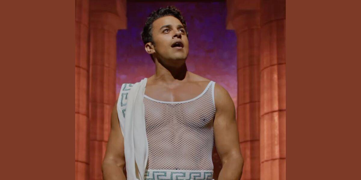 A man wearing a white mesh top and a toga-style garment stands in front of large columns with a surprised expression, against a purple and red background.