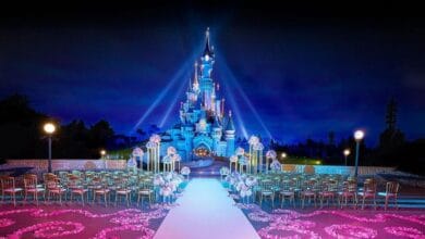 A nighttime Disneyland Paris wedding setup in front of a brightly lit fairytale castle, with rows of golden chairs and a white aisle surrounded by pink and purple flower arrangements. Blue spotlights illuminate the enchanting scene.