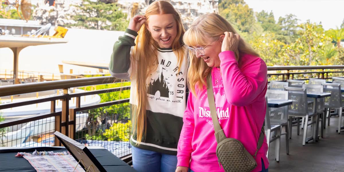 Two women smiling and laughing while looking at items on a table outdoors. Both are wearing Disneyland spirit jerseys, and one has a crossbody bag. Trees and tables are visible in the background.