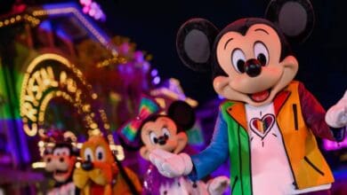 Mickey Mouse sports a colorful jacket with a heart design, smiling in front of Minnie, Goofy, and Pluto at a brightly lit, festive parade—showcasing Disney pride merch during a joyful night celebration.