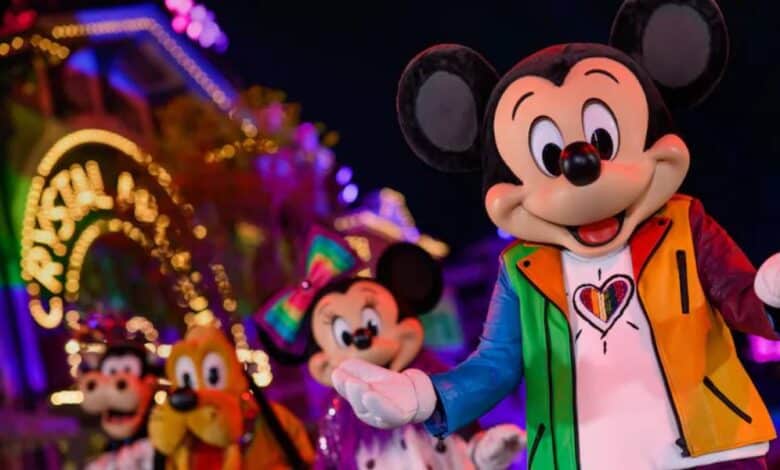 Mickey Mouse sports a colorful jacket with a heart design, smiling in front of Minnie, Goofy, and Pluto at a brightly lit, festive parade—showcasing Disney pride merch during a joyful night celebration.