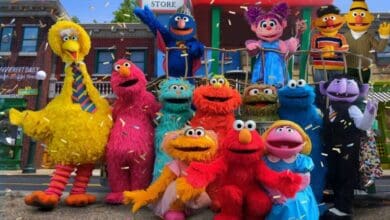 A colorful group of Sesame Street puppet characters, including Big Bird, Elmo, Cookie Monster, and others, pose and smile together outdoors on a city street, confetti falling around them.