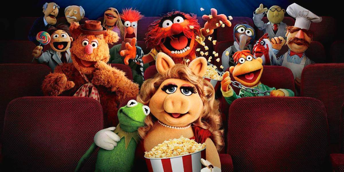 The Muppets sit in a movie theater, smiling and excited. Miss Piggy and Kermit are in the front with popcorn, surrounded by other colorful Muppet characters filling the red seats.
