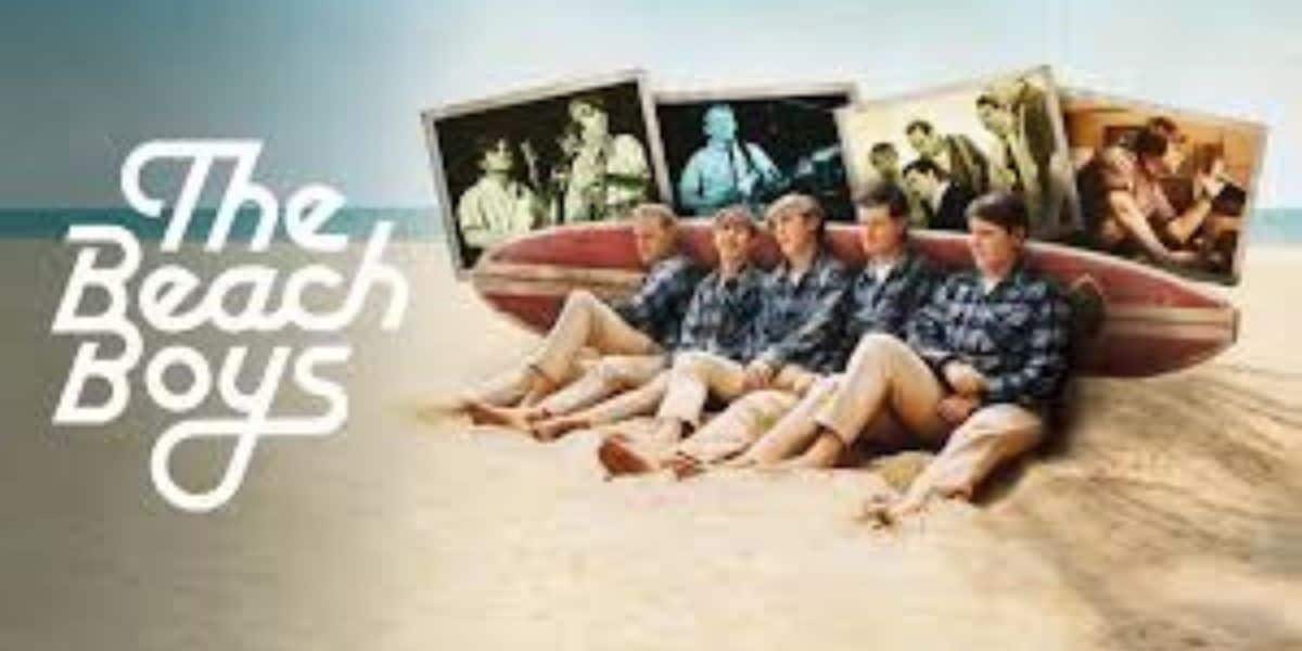 Five men wearing matching shirts sit on a sandy beach in front of a striped surfboard. "The Beach Boys" is written in white text, with vintage band photos displayed above the group.