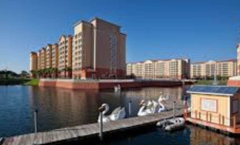 Several tall, modern apartment buildings line the edge of a lake with a dock in the foreground. Swan-shaped pedal boats and a small wooden structure are visible by the dock under a clear blue sky.