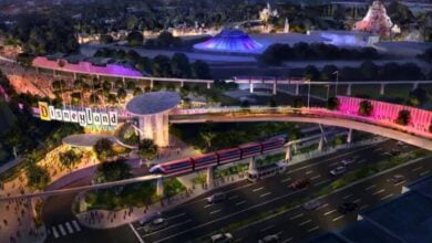 A colorful, futuristic rendering of Disney’s Disneyland entrance featuring the iconic sign, a monorail gliding above a busy road, crowds of people, and theme park attractions in the background at dusk.
