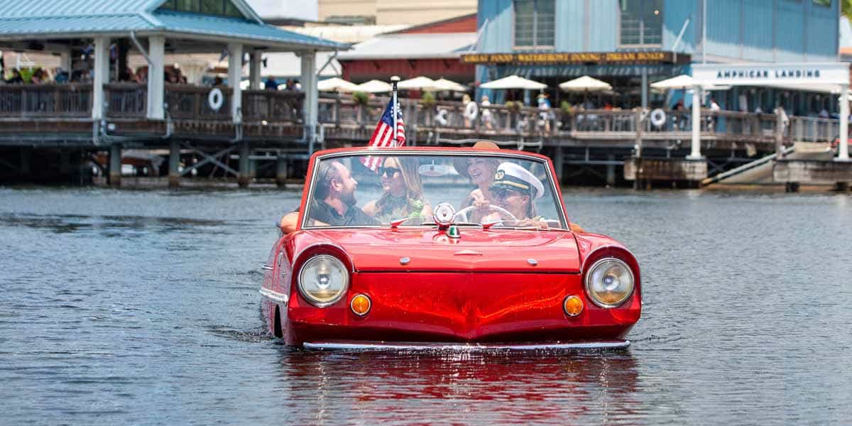 A red Disney Springs Amphicar Tours vehicle with people inside drives on water near a waterfront area with restaurants and docks. An American flag is attached to the back of the car.