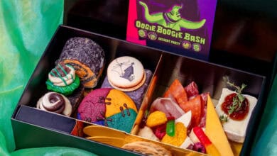 A black box labeled "Oogie Boogie Bash Dessert Party" contains colorful cookies, rolled pastries, candy, cheese, crackers, salami, and a decorated macaron with a Halloween theme. The box sits on green fabric.