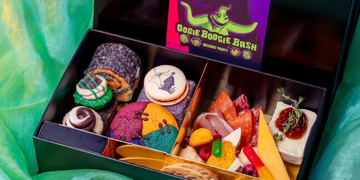 A black box labeled "Oogie Boogie Bash Dessert Party" contains colorful cookies, rolled pastries, candy, cheese, crackers, salami, and a decorated macaron with a Halloween theme. The box sits on green fabric.