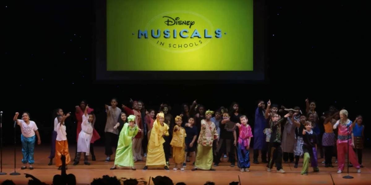 A large group of children in colorful costumes perform on stage beneath a projected Disney Musicals in Schools logo, striking lively poses as they sing and dance.