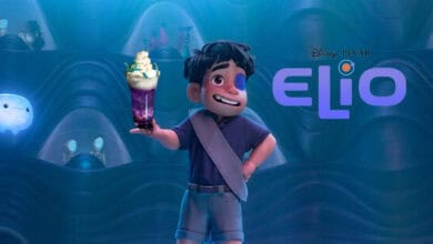 A boy with dark hair and a blue shirt stands smiling and holding a colorful drink with whipped cream. He is in a blue, futuristic room with glowing orbs. The Disney Pixar "Elio" logo appears beside him.