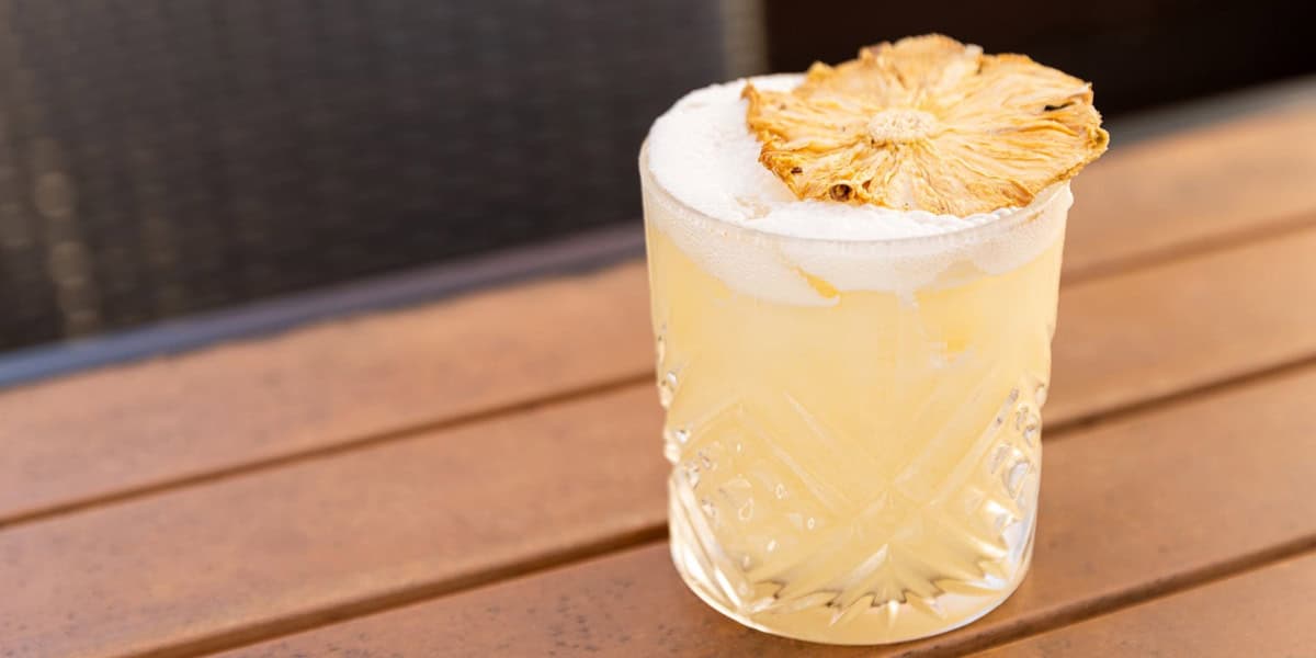 A cocktail in a textured glass on a wooden table, garnished with a dried pineapple slice and a foamy top.