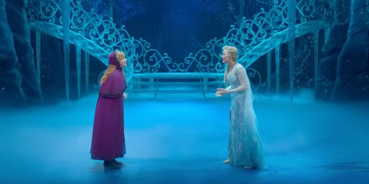 Two women in costume stand facing each other on a blue-lit stage with icy, ornate gates and a bench in the background, resembling a scene from Disney’s Frozen musical adaptation.