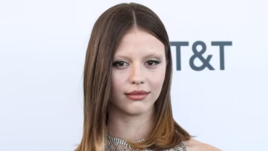 A woman with straight, shoulder-length hair wearing a black dress with a sparkly, netted neckline poses in front of a white background featuring partial logos and text, echoing the excitement of Mia Goth cast in Star Wars.