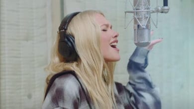 A woman with long blonde hair wearing headphones sings passionately into a studio microphone, holding one hand up, in a recording booth with wooden walls.