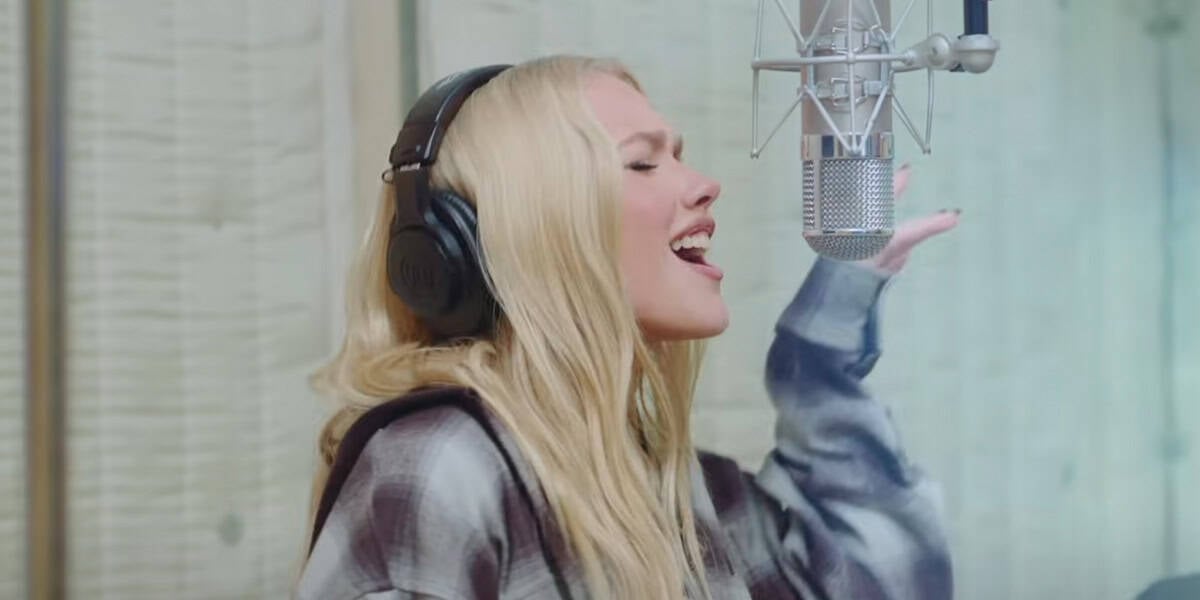A woman with long blonde hair wearing headphones sings passionately into a studio microphone, holding one hand up, in a recording booth with wooden walls.