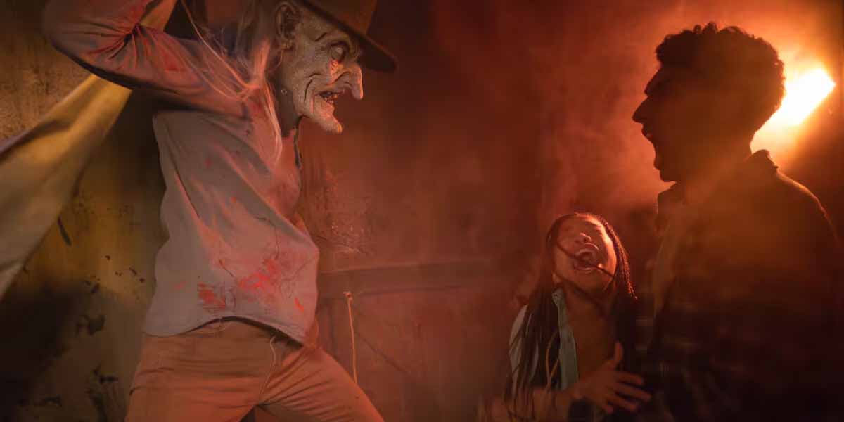 A creepy figure in a hat and tattered clothes looms menacingly over two terrified people, a man and a woman, in a dimly lit, orange-toned room. The woman clings to the man, both screaming in fear.