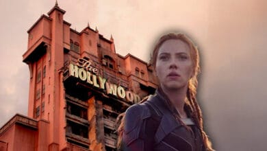 A woman in a black costume stands in front of the Hollywood Tower Hotel, a tall, reddish building with a vintage sign, under a cloudy sky.