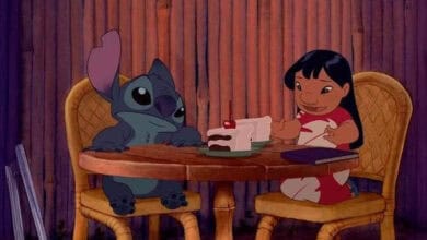 Animated characters Stitch and Lilo sit at a round wooden table with slices of cake and a book, enjoying Stitch-inspired dining for 626 day. Stitch eyes the cake while Lilo smiles, set against a wooden backdrop and wicker chairs.