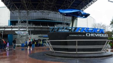 People walk near the Test Track attraction entrance at Epcot, featuring a large sign with the Chevrolet logo and a blue car model above it under a metal canopy on a wet day.