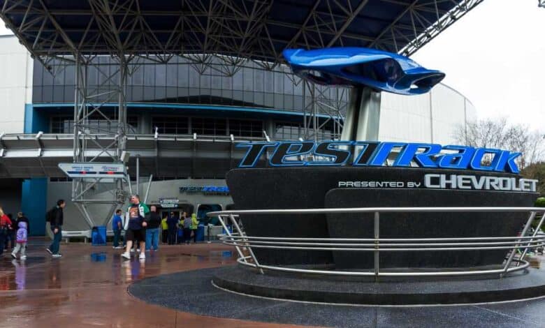 People walk near the Test Track attraction entrance at Epcot, featuring a large sign with the Chevrolet logo and a blue car model above it under a metal canopy on a wet day.