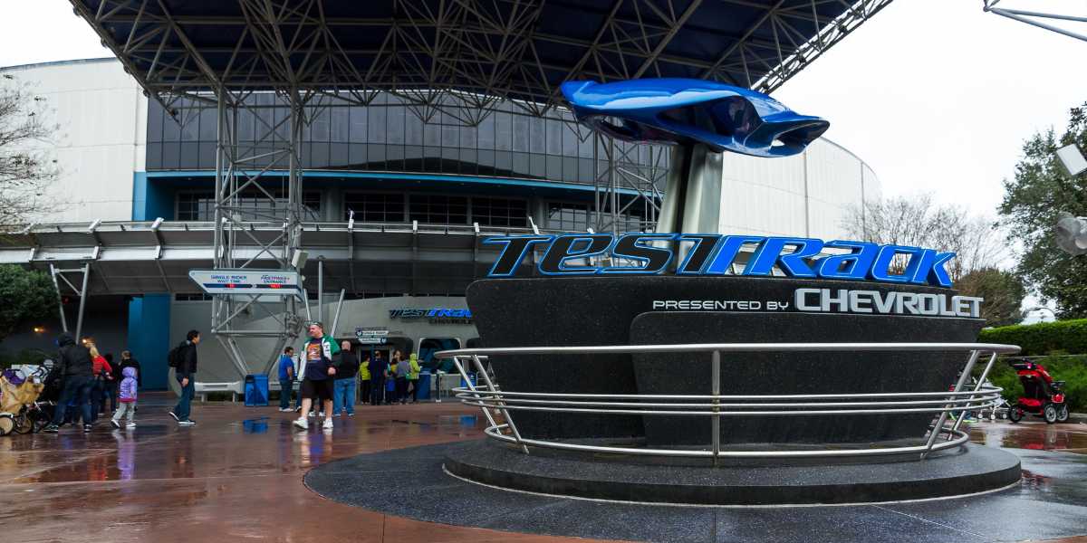 People walk near the Test Track attraction entrance at Epcot, featuring a large sign with the Chevrolet logo and a blue car model above it under a metal canopy on a wet day.