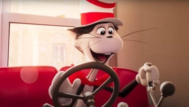 Animated Cat in the Hat character, wearing a red and white striped hat and red bow tie, sits in a red car, smiling and holding the steering wheel and gearshift with a window in the background.
