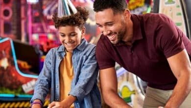 A smiling child in a party hat and a man play an arcade game together, enjoying themselves in a brightly lit amusement arcade.