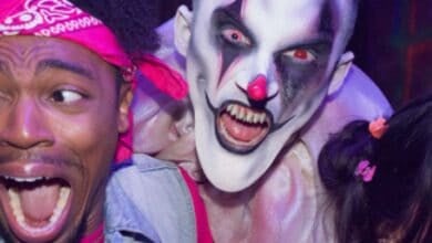 A person in clown makeup with sharp teeth and red eyes screams behind a frightened man wearing a pink bandana at Fright Fest Six Flags. Another person is partially visible on the right in this intense, scary scene.