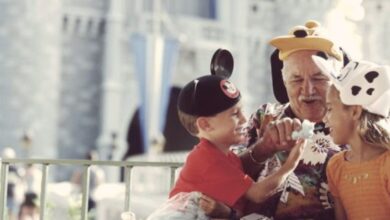 An elderly man and two children wearing Disney hats laugh and share a treat in front of a castle at a theme park, likely Disneyland or Disney World. The atmosphere is joyful and playful.