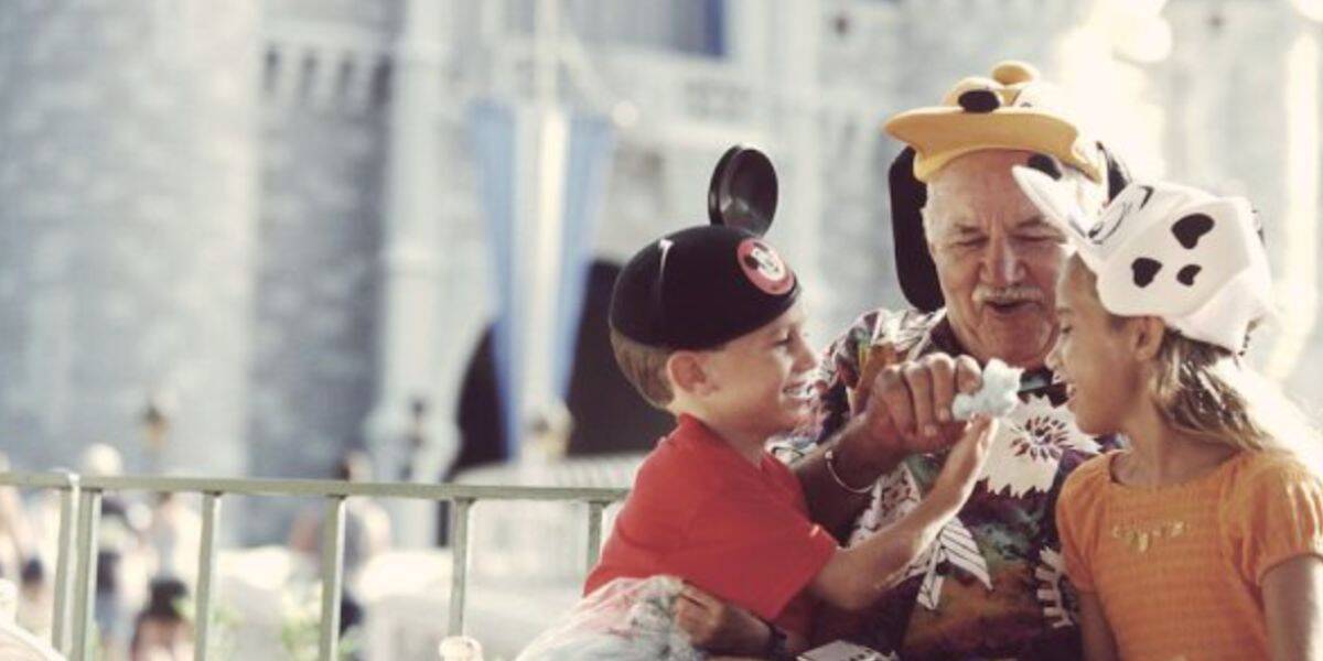 An elderly man and two children wearing Disney hats laugh and share a treat in front of a castle at a theme park, likely Disneyland or Disney World. The atmosphere is joyful and playful.