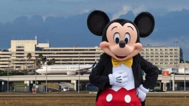 Mickey Mouse costume stands in front of an airport terminal, with an airplane and the building visible in the background MCO.
