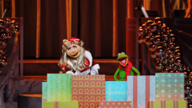 Miss Piggy and Kermit the Frog stand behind colorful wrapped presents on stage, decorated with garlands and lights. Miss Piggy wears a Santa hat and red dress, while Kermit sports a red scarf.