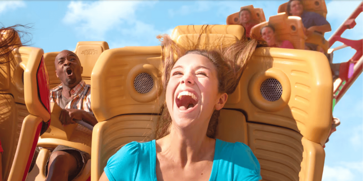 A woman in a teal shirt laughs with her hair flying up while riding a roller coaster, with other riders behind her showing excitement and surprise against a blue sky.