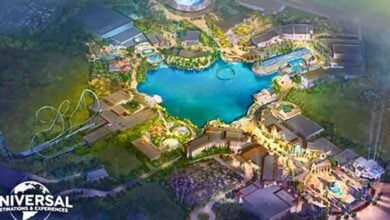 A colorful aerial illustration of a theme park with a large central lake, roller coasters, attractions, and buildings, featuring the Universal Destinations & Experiences logo in the bottom left corner.