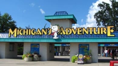 Entrance to Michigan’s Adventure amusement park, featuring a large yellow sign with Snoopy in aviator gear. Below the sign are ticket booths, colorful flowers, and a few people at the entrance. This Six Flags theme park is rumored for a closure.