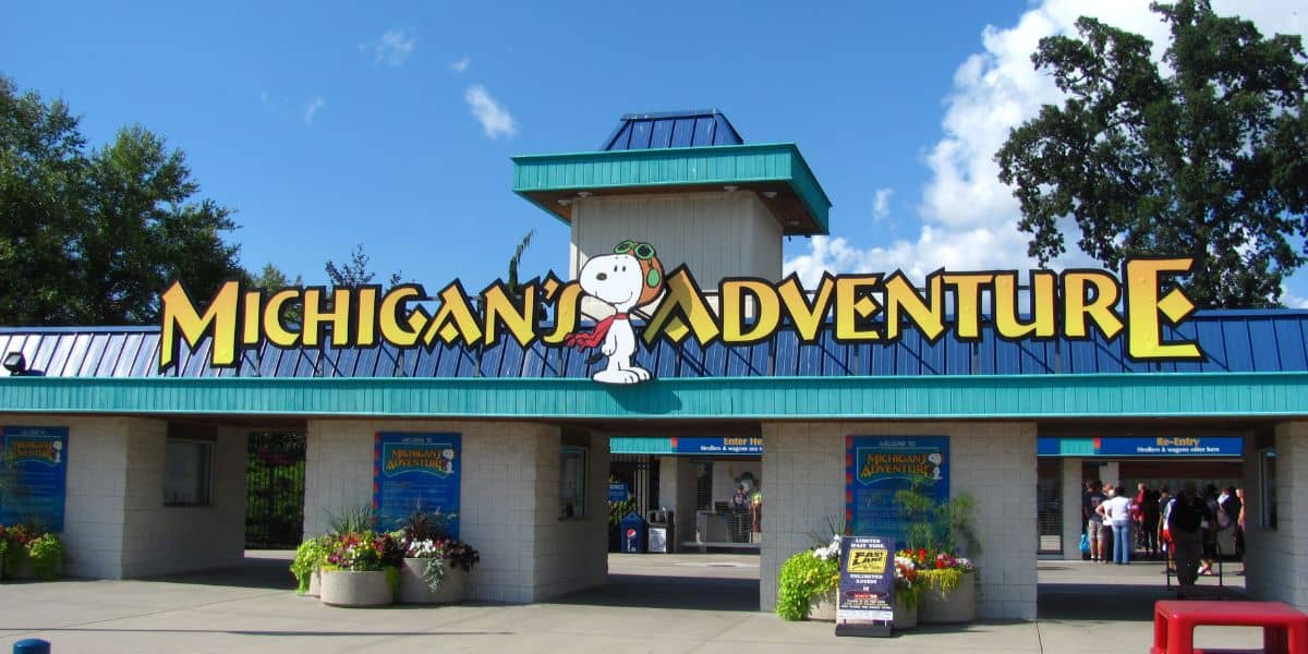 Entrance to Michigan’s Adventure amusement park, featuring a large yellow sign with Snoopy in aviator gear. Below the sign are ticket booths, colorful flowers, and a few people at the entrance. This Six Flags theme park is rumored for a closure.
