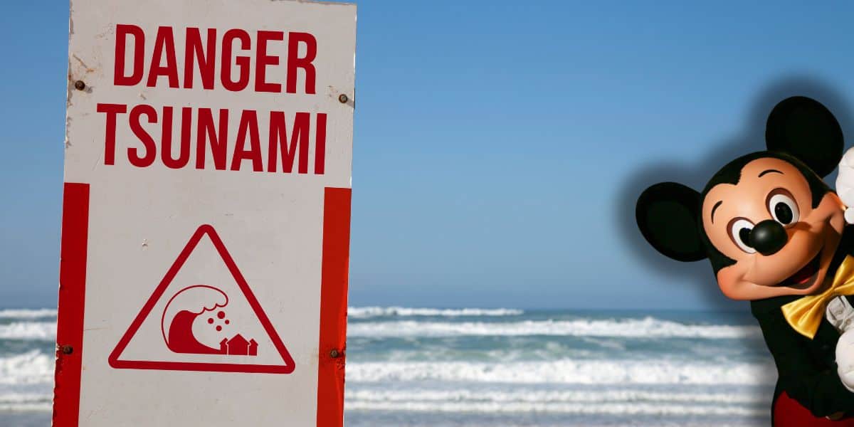 A "Danger Tsunami" warning sign stands on a beach with waves in the background. On the right, a person in a Mickey Mouse costume is peeking playfully into the frame as Disney locations deal with tsunami waves and warning.