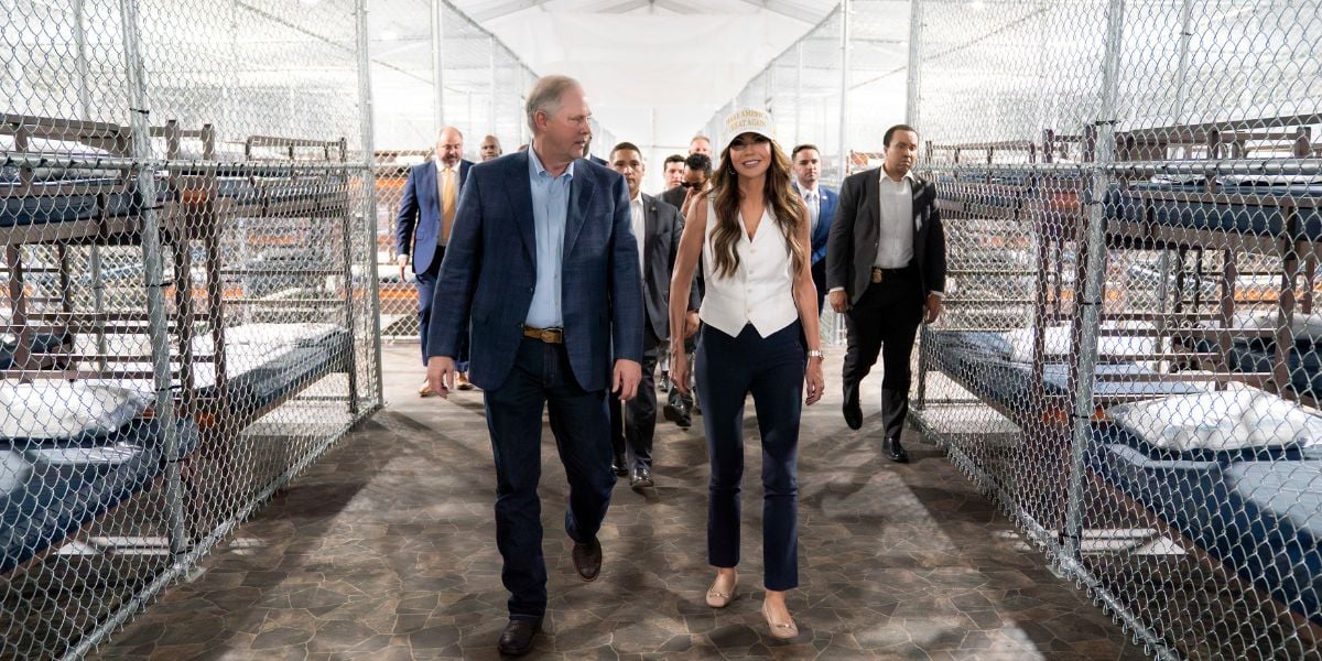 A group of people walk through a facility with chain-link fenced enclosures containing bunk beds. Two people lead the group, talking, while others follow behind. The area is brightly lit and has a tiled floor.