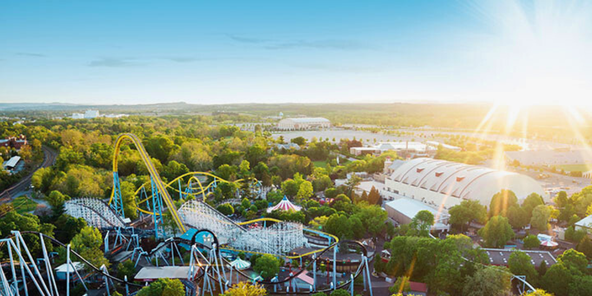 A bright, sunny view of an amusement park featuring roller coasters, trees, and large white domed buildings, with green landscape and distant hills under a clear blue sky, where tragic theme park news is given.