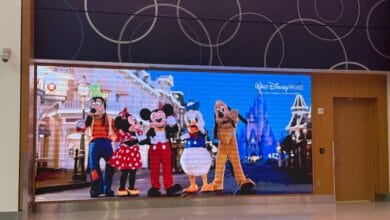 A digital display outside the Disney store at MCO shows Goofy, Minnie Mouse, Mickey Mouse, Donald Duck, and Pluto posing together on Main Street with Cinderella Castle in the background. The display advertises Walt Disney World.