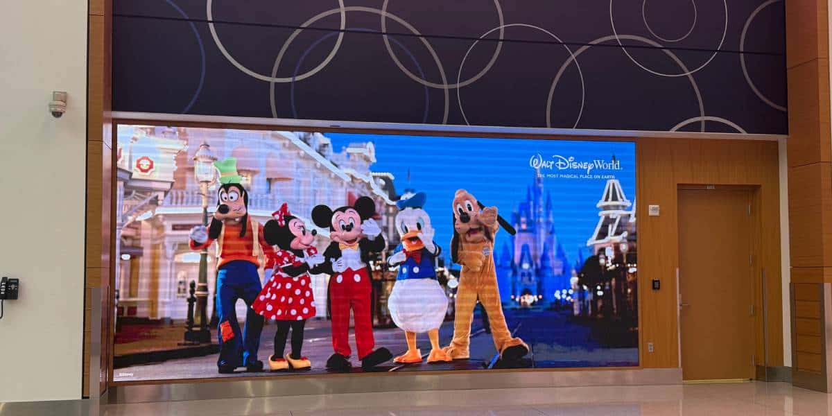 A digital display outside the Disney store at MCO shows Goofy, Minnie Mouse, Mickey Mouse, Donald Duck, and Pluto posing together on Main Street with Cinderella Castle in the background. The display advertises Walt Disney World.
