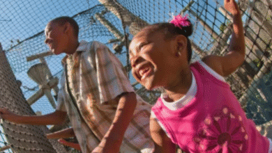 Two children, a boy and a girl, smile and play outside near a tall wire fence on a sunny day, reminiscent of Jurassic Park. The girl wears a pink shirt and hair ties; the boy wears a plaid shirt. Trees and blue sky fill the background.