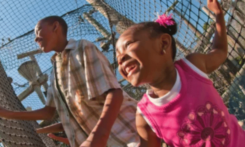 Two children, a boy and a girl, smile and play outside near a tall wire fence on a sunny day, reminiscent of Jurassic Park. The girl wears a pink shirt and hair ties; the boy wears a plaid shirt. Trees and blue sky fill the background.