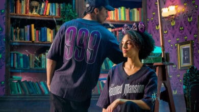 Two people in a spooky, purple-lit library sport Haunted Mansion 2025 merch jerseys—one with "999" on the back, the other with "Haunted Mansion" on the front. The woman also wears sparkly cat ears. Green eyes decorate the wallpaper.